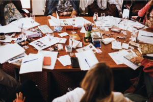A cluttered table with people out of view sitting around it. papers pens and galsses of water.